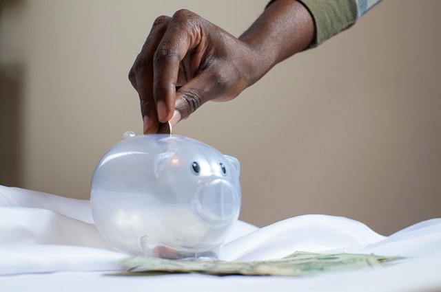 Photo of a black hand putting coins inside of a clear piggybank