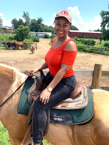 A light skinned black woman smiles and poses for a photo atop her camel-coloured horse. She is earring a beige and red cap, black jeans and a red shirt.