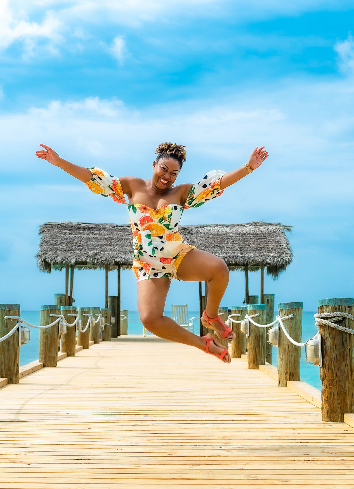 This Bahamian Gyal blogger, Rogan Smith jumps in the air with her hands outstretched. She is on a dock with the gorgeous blue ocean and sky behind her