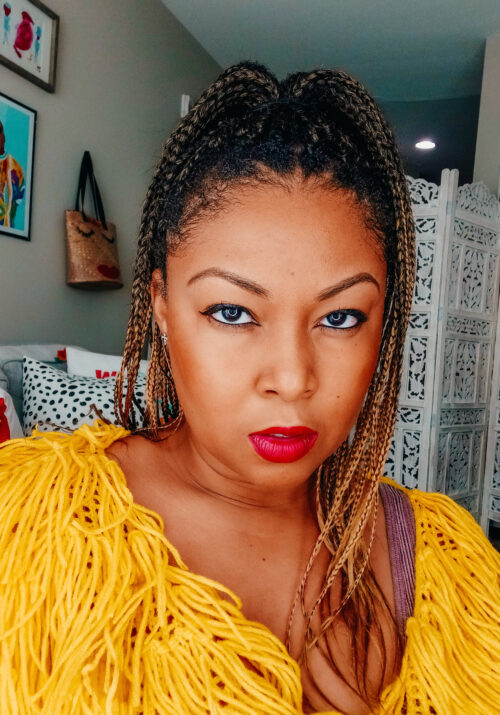 Blogger Rogan wears honey coloured box braids in her hair. She is wearing a vibrant yellow yarn jacket and stares directly into the camera.