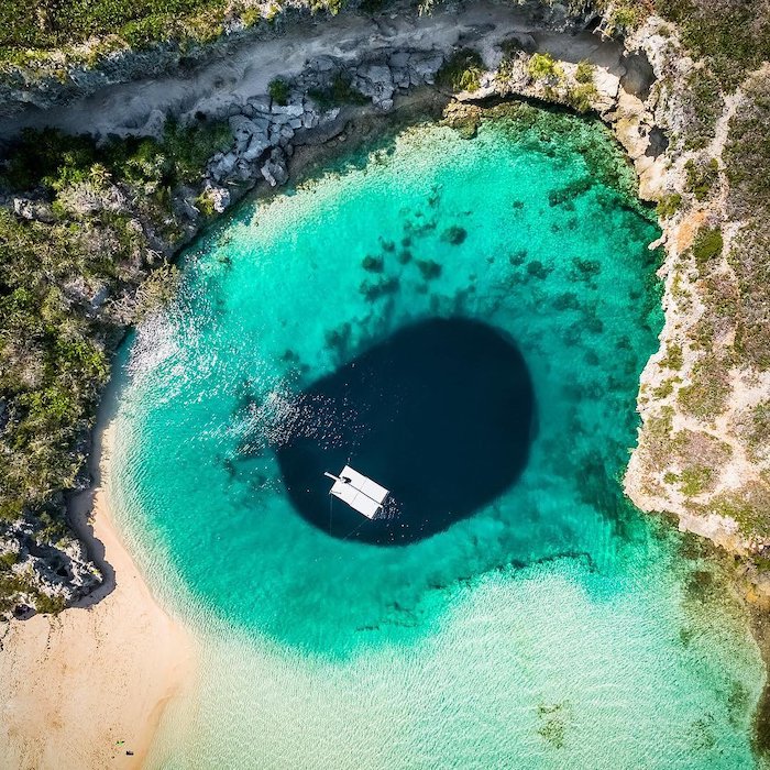 Aerial image of Dean's Blue Hole in Long Island, Bahamas. A deep dark blue hole is surrounded by aquamarine waters on a white sandy beach. Here's a Bahamas fun fact: it's the second deepest blue hole in the world. 