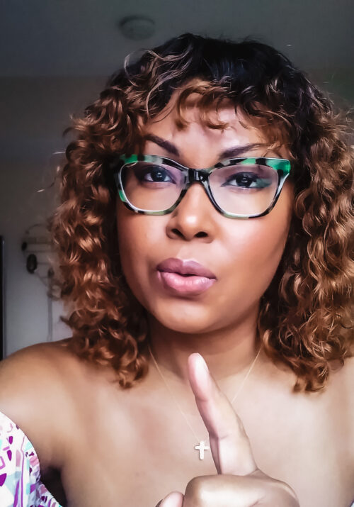 Bahamian writer, Rogan Smith wears green tortoise glasses and curly hair. She points her finger at camera.