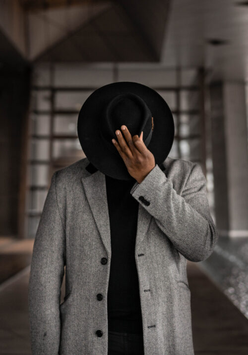 Photo shows a black man in a chic grey jacket and black shirt hiding his face with a fedora. Photo by Luca Nardone