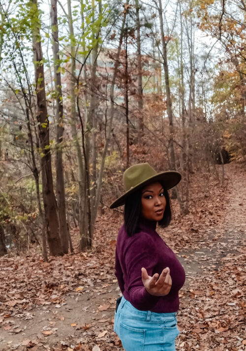 A black woman wearing a green fedora and a purple shirt walks into the woods. She has a smirk on her face and gestures to the camera to come long.