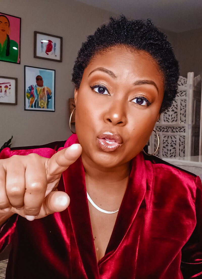 A light-skinned black woman wearing a red velvet blazer points at the camera in a condemning way. She has a serious expression on her face.