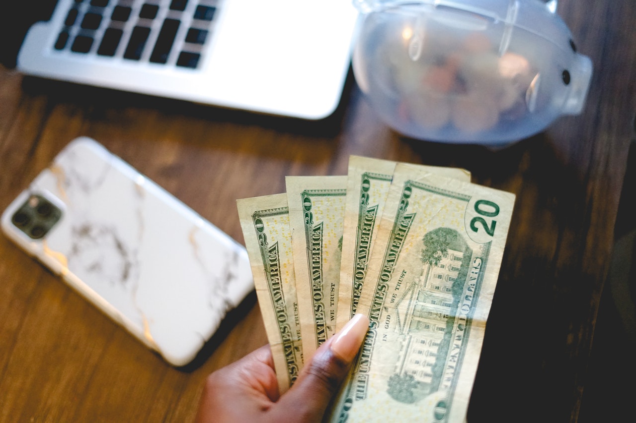 A Black woman's hand is shown holding money. A computer sits in the background.