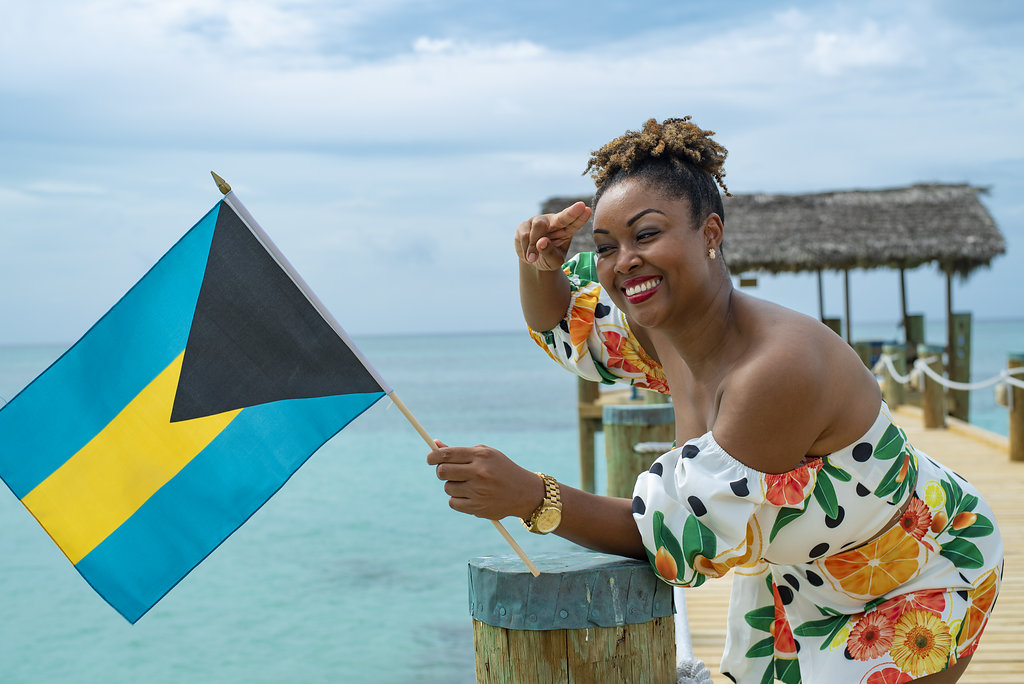 Bahamian Blogger Rogan Smith poses on a beach with a Bahamian flag in her hand. She salutes the flag with her right hand