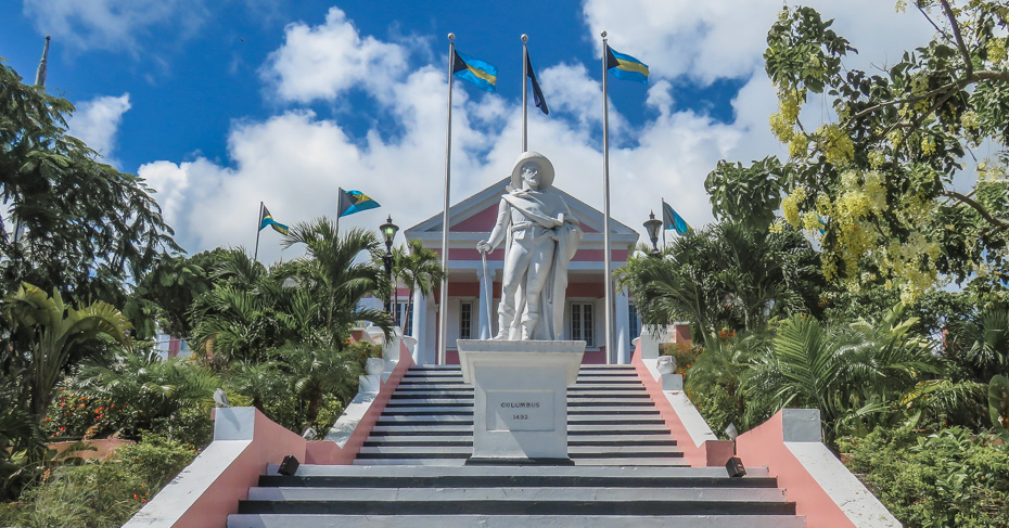 A Christopher Columbus statue stands in front of Government House in The Bahamas. Several Bahamian flags fly in the background and trees surround the statue.  