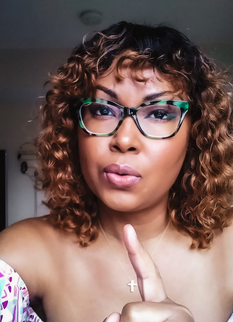 Bahamian writer, Rogan Smith wears green tortoise glasses and curly hair. She points her finger at camera.