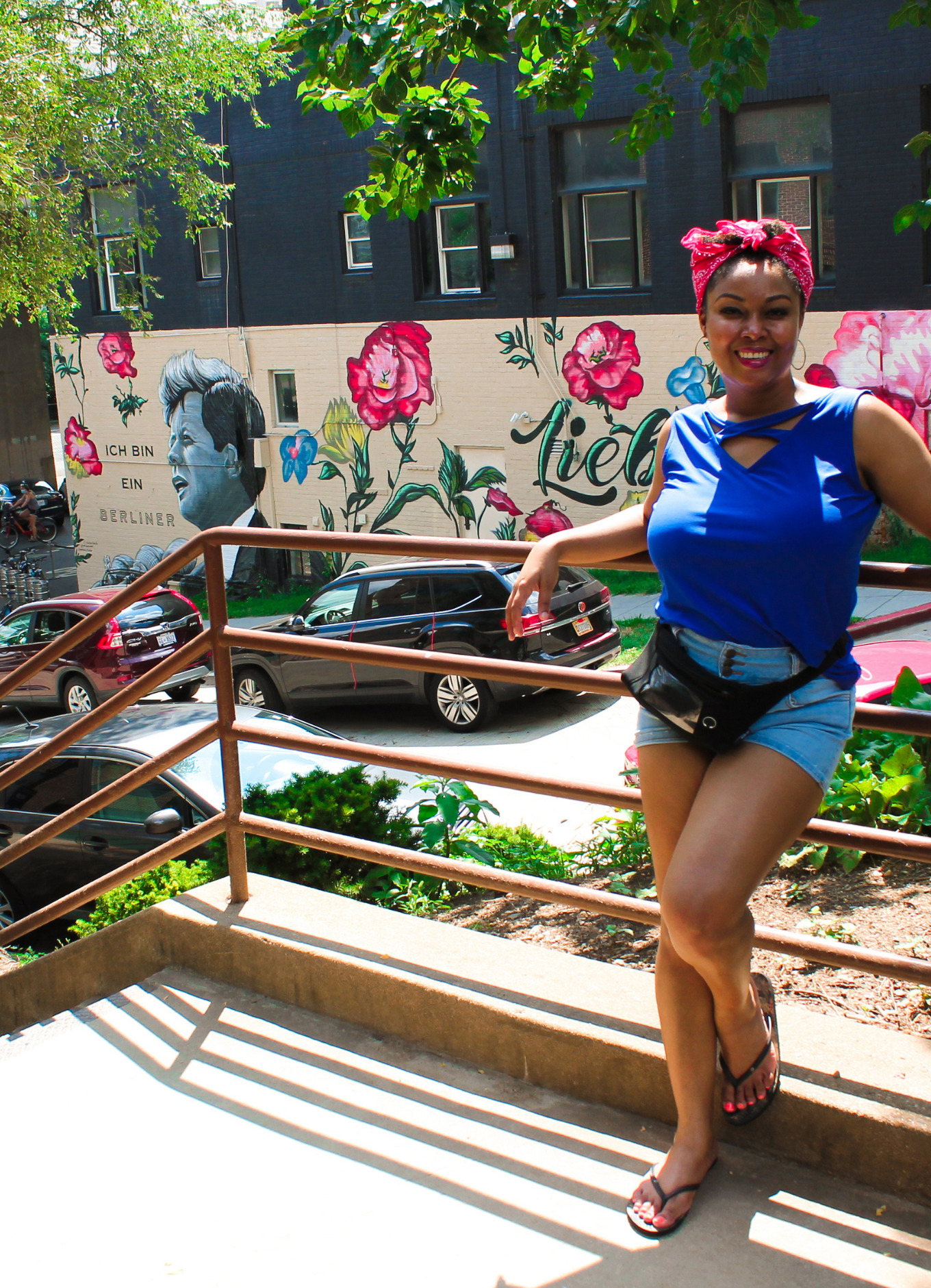 Blogger Rogan Smith wears a red bandana and a blue shirt. She poses against a rail with a mural of John F Kennedy in the background