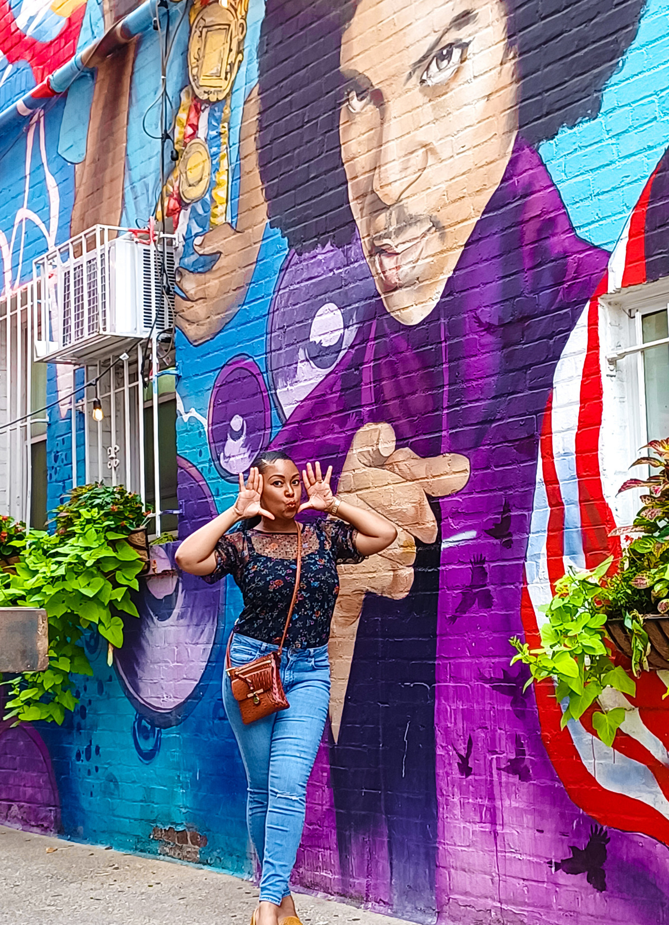 Rogan smiles and poses in front of a purple Prince mural next to Ben's Chili Bowl in northwest Washington, DC. She is wearing blue denim pants and a flower blouse.