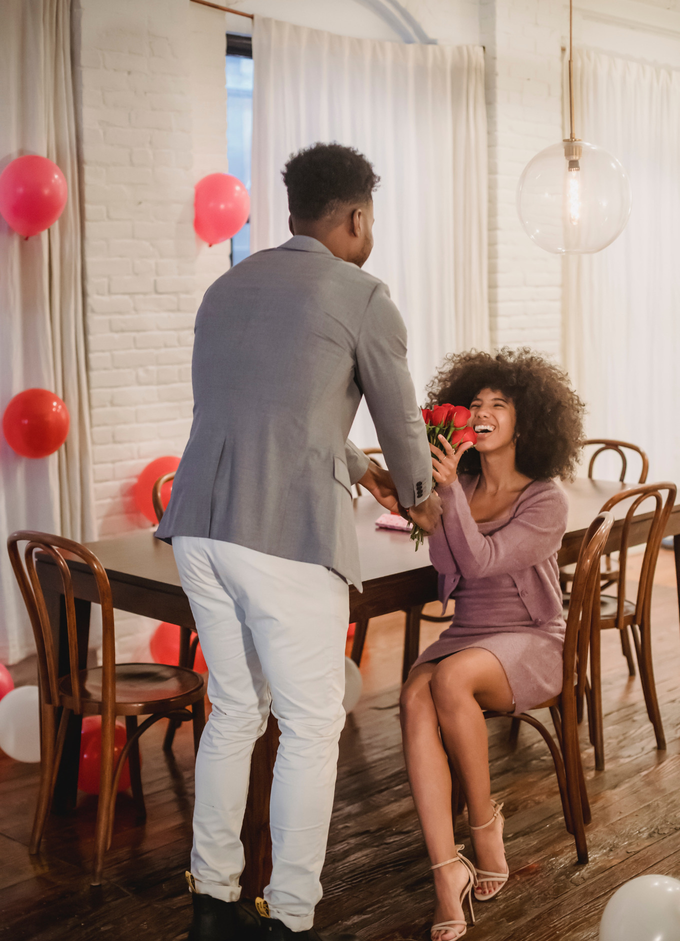 A black man in a gray jacket and white pants gives red roses to a black woman with big, curly hair