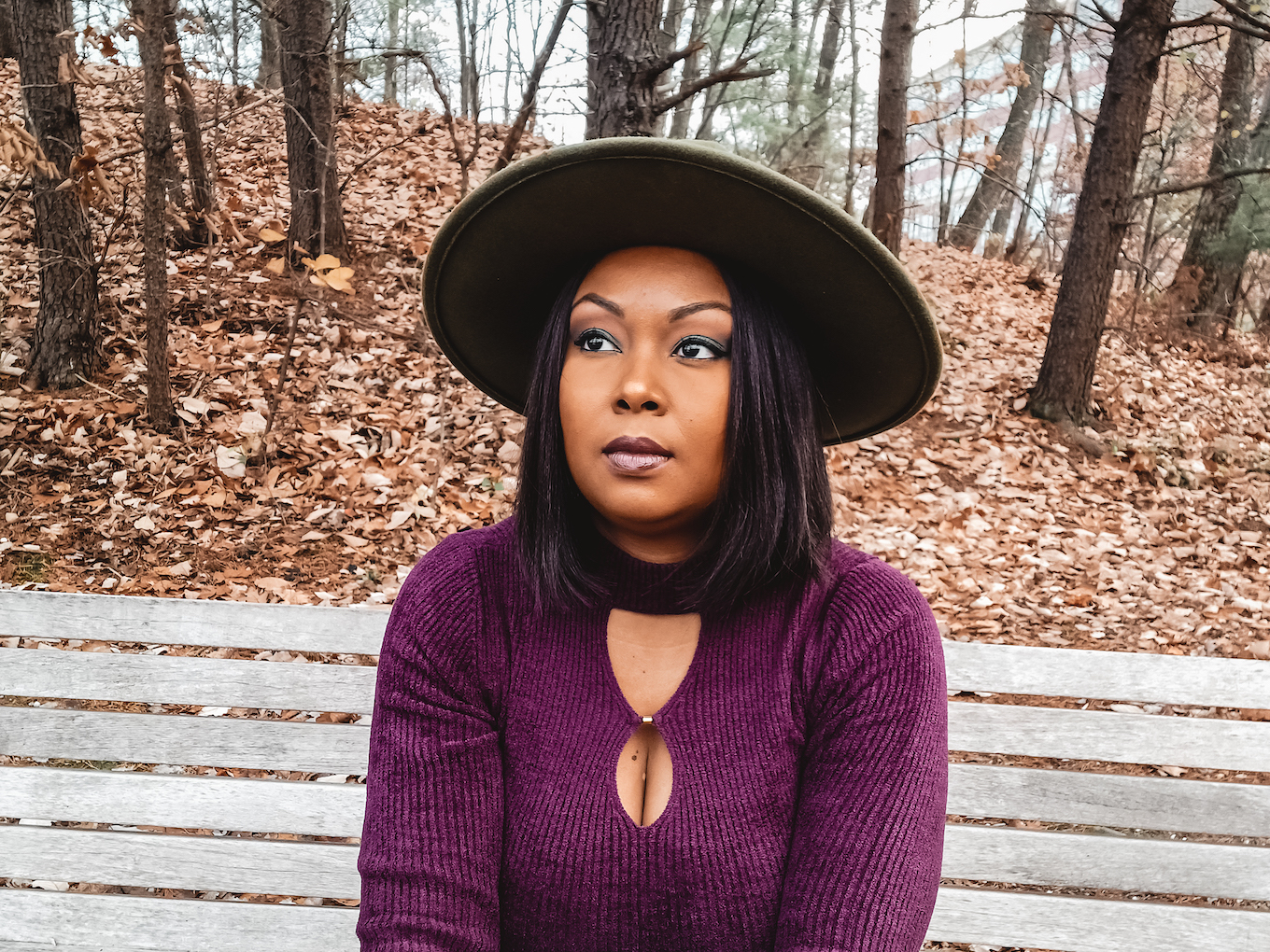 This Bahamian Gyal blogger Rogan sits on a bench in the woods. She wears a plum coloured shirt and a green fedora. She sits on a bench on a wooded trail.
