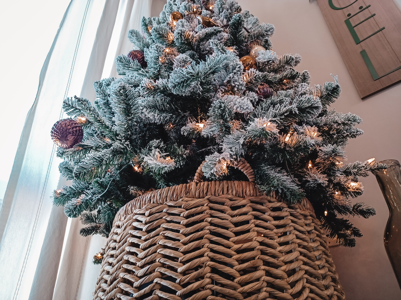 A tall flocked Christmas tree sits snugly inside a tan-coloured wicker basket. 