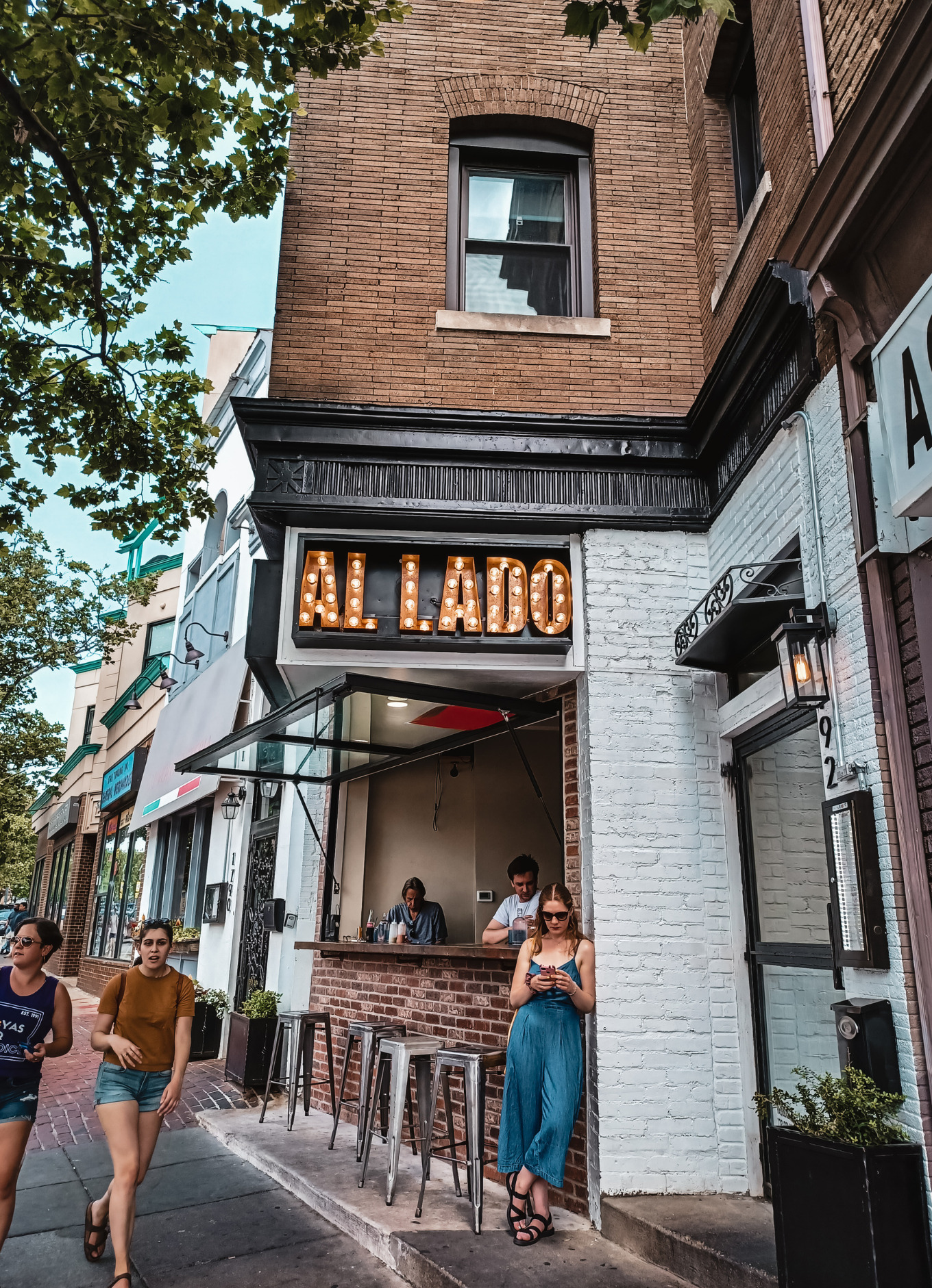 A white woman leans on the side of Al Lado restaurant while looking at her cell phone. Two women walk by talking as they take in the sights of Adams Morgan, one of the most walkable areas in Washington, DC In the Washington, DC vs Virginia battle for walkability, DC wins. 