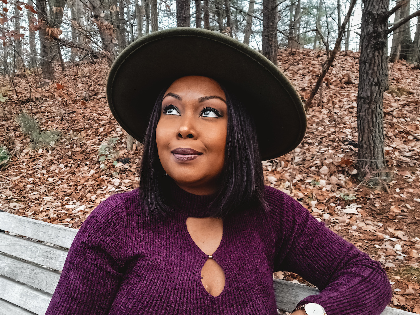 This Bahamian Gyal blogger Rogan smith smiles as she sits on an empty bench in the woods in Virginia. She is wearing a plum shirt and a green fedora. She takes in the natural beauty of the surrounding area.