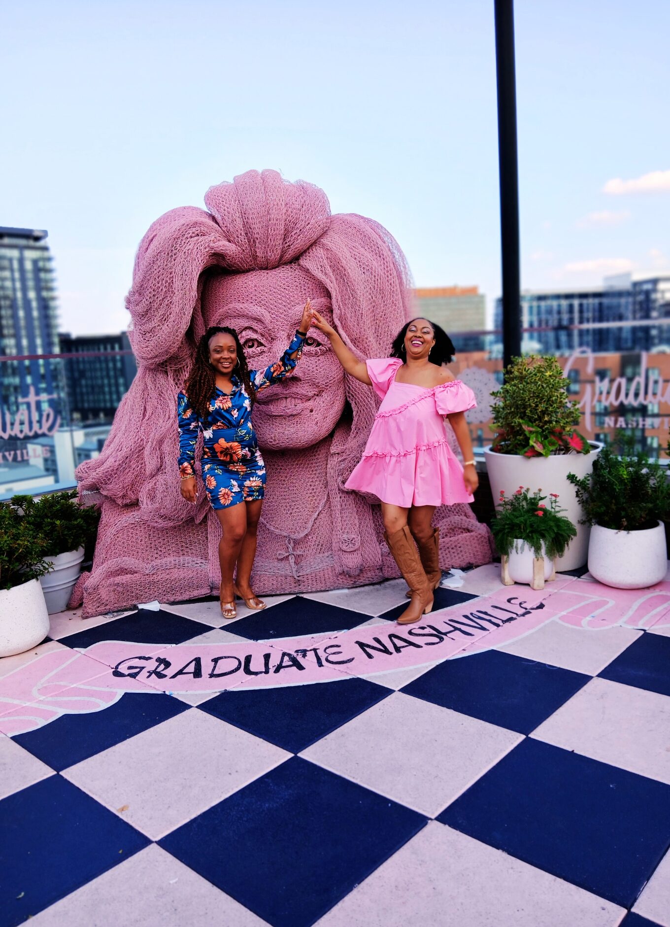 This Bahamian Gyal blogger, Rogan wears a pink babydoll dress and brown cowboy boots. She and her friend, who is wearing blue, smile and pose in front of the Dolly Parton sculpture at White Limozeen cocktail bar.