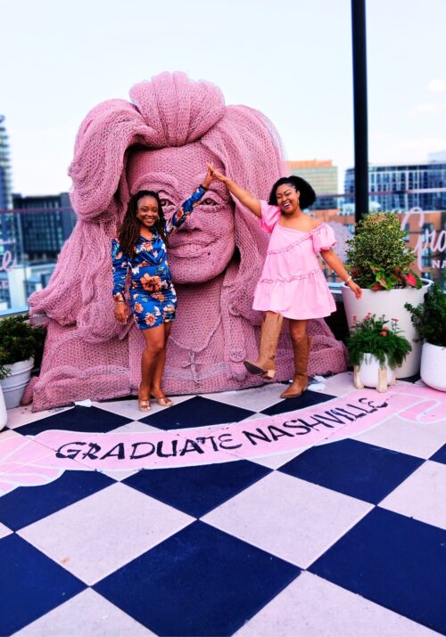 This Bahamian Gyal blogger, Rogan wears a pink babydoll dress and brown cowboy boots. She and her friend, who is wearing blue, smile and pose in front of the Dolly Parton sculpture at White Limozeen cocktail bar.