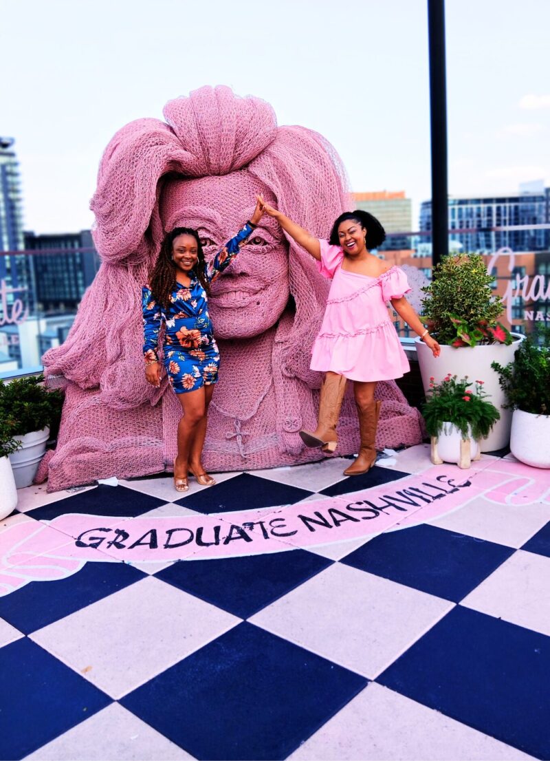 This Bahamian Gyal blogger, Rogan wears a pink babydoll dress and brown cowboy boots. She and her friend, who is wearing blue, smile and pose in front of the Dolly Parton sculpture at White Limozeen cocktail bar.