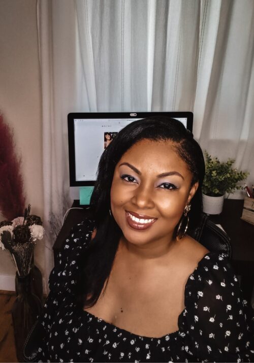 This Bahamian Gyal blogger, Rogan sits in front of a MAC computer screen and a tidy desk. She is wearing a black flowy blouse and her long hair hangs over her shoulder.