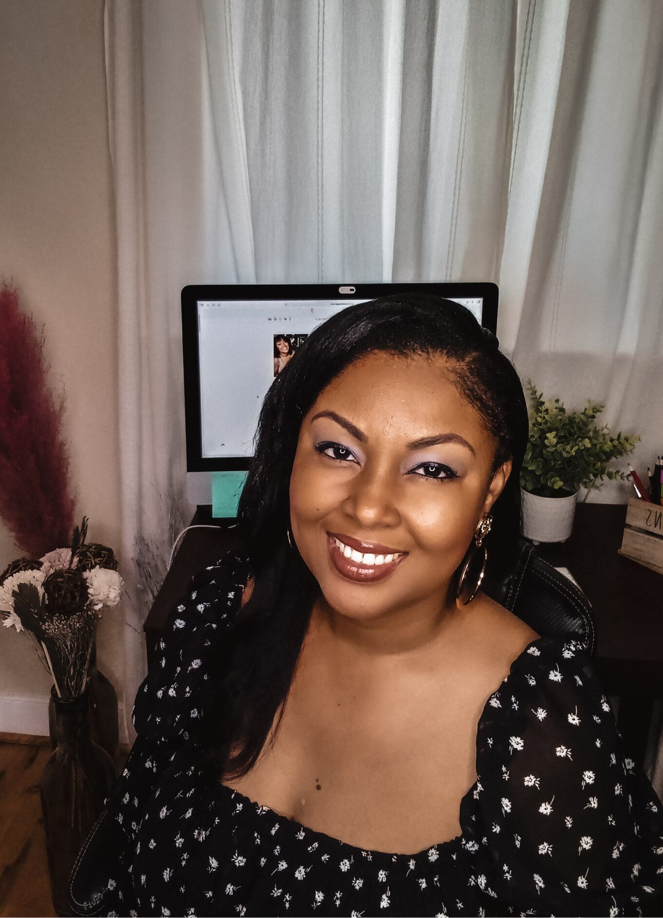 This Bahamian Gyal blogger, Rogan sits in front of her MAC computer screen working from home. Her website's landing page can be seen on her screen. She is wearing a black flowy top and her long hair hangs over her shoulder.