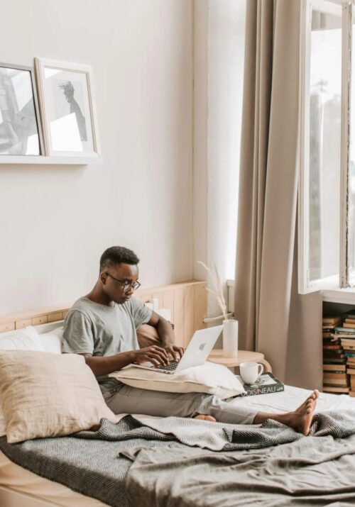 A black man sits on bed on Apple laptop as he works from home