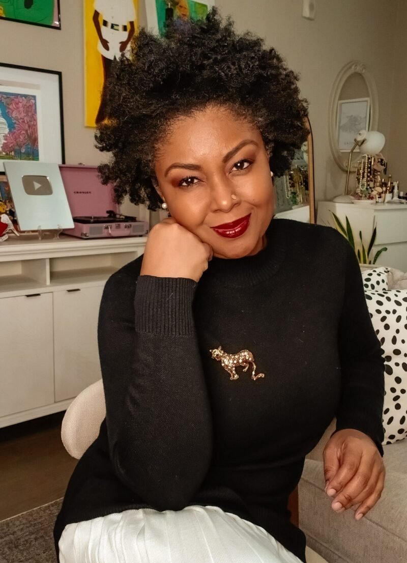 How To Cultivate Gratitude On Thanksgiving - This Bahamian Gyal blogger, Rogan sits in her living room. She is sporting a curly afro, a black sweater, a pleated cream skirt and a jaguar pin