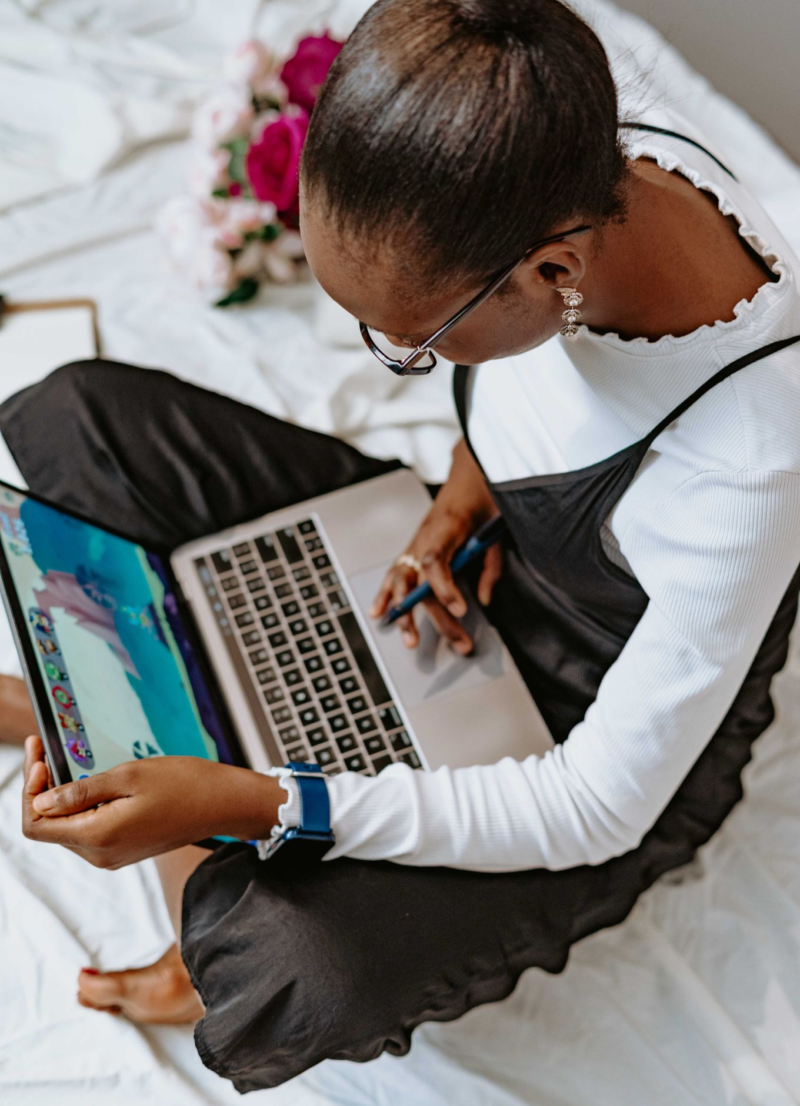 An African American woman sits on a bed with her Apple laptop in hand, reading an email about a YouTube sponsorship scam.